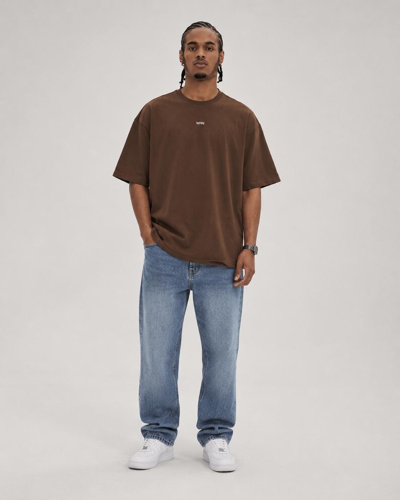 May include: A person wearing a brown t-shirt with a small logo and blue jeans. The t-shirt is a solid color and has short sleeves. The jeans are a light blue wash and have a relaxed fit. White sneakers complete the outfit.
