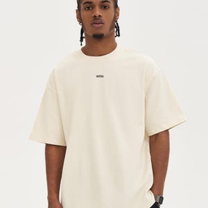 May include: A cream-coloured, short-sleeved t-shirt with the word "WRN" printed in black on the chest. The shirt has a relaxed fit and a crew neck. The model is wearing black trousers and a watch.