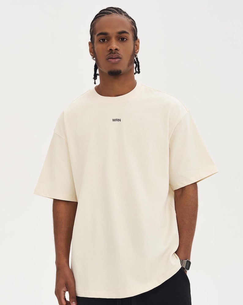 May include: A cream-coloured, short-sleeved t-shirt with the word "WRN" printed in black on the chest. The shirt has a relaxed fit and a crew neck. The model is wearing black trousers and a watch.