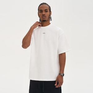 May include: A white, short-sleeved t-shirt with the word "WRN" printed in black on the chest. The shirt has a crew neck and a relaxed fit. The person in the image is wearing black pants and a silver watch.