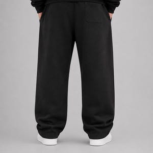 Puede incluir: Pantalones de ch&aacute;ndal negros lisos con un corte holgado y ancho, cintura el&aacute;stica gruesa y un bolsillo de parche en la parte trasera derecha. Los pantalones se combinan con zapatillas blancas, mostrando un dise&ntilde;o limpio y sencillo.