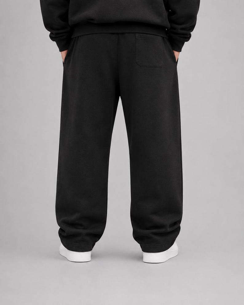 Puede incluir: Pantalones de ch&aacute;ndal negros lisos con un corte holgado y ancho, cintura el&aacute;stica gruesa y un bolsillo de parche en la parte trasera derecha. Los pantalones se combinan con zapatillas blancas, mostrando un dise&ntilde;o limpio y sencillo.