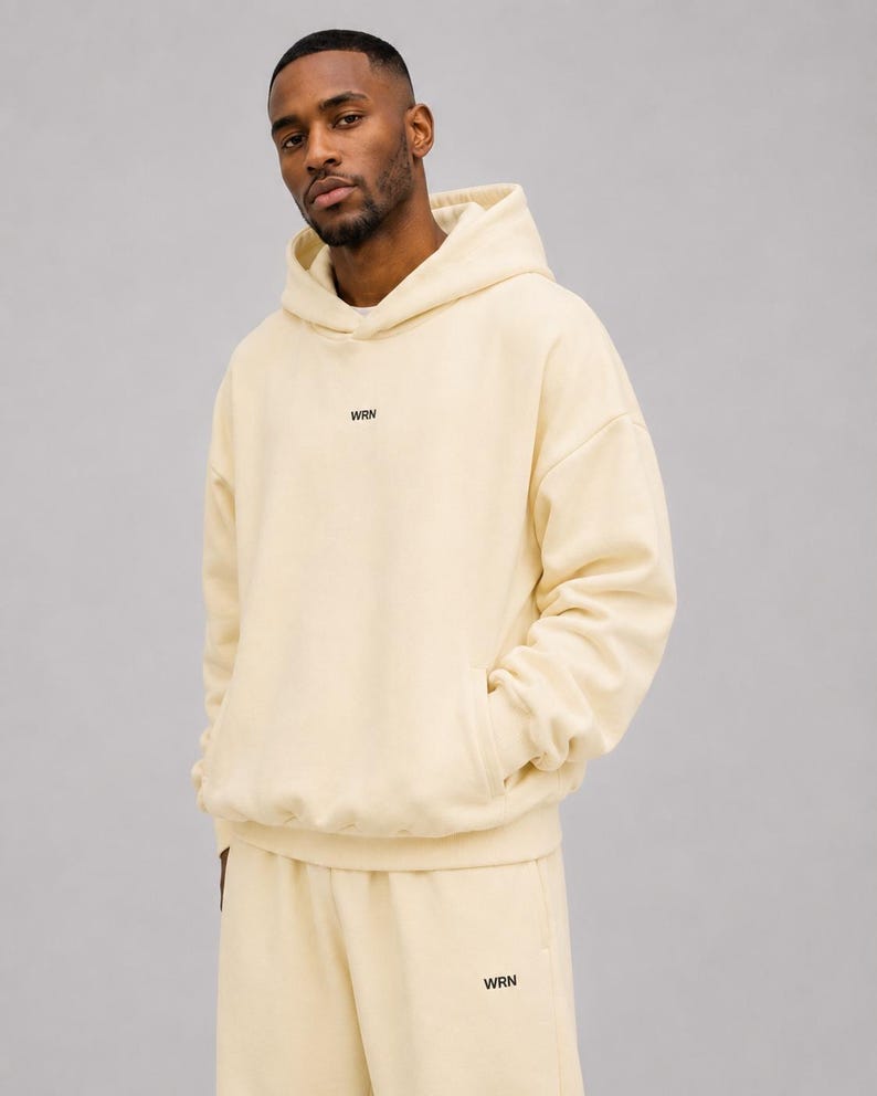 May include: A cream-coloured hooded sweatshirt featuring a front kangaroo pocket and the letters WRN printed in small black font on the centre of the chest. The matching cream-coloured sweatpants also feature the letters WRN in black on the leg.