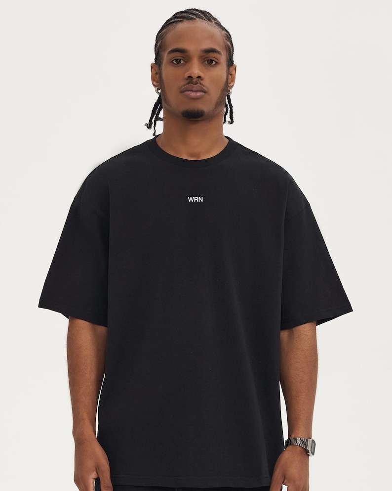 May include: Black short-sleeved t-shirt with the letters "WRN" printed in white on the chest. The shirt has a relaxed fit and a crew neck. The model is wearing a silver watch.