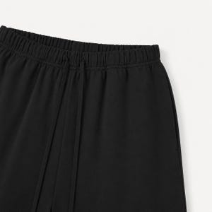 May include: Close-up of black trousers featuring an elasticated waistband and a drawstring. The material looks smooth and opaque. These trousers are likely part of a casual or sportswear collection.