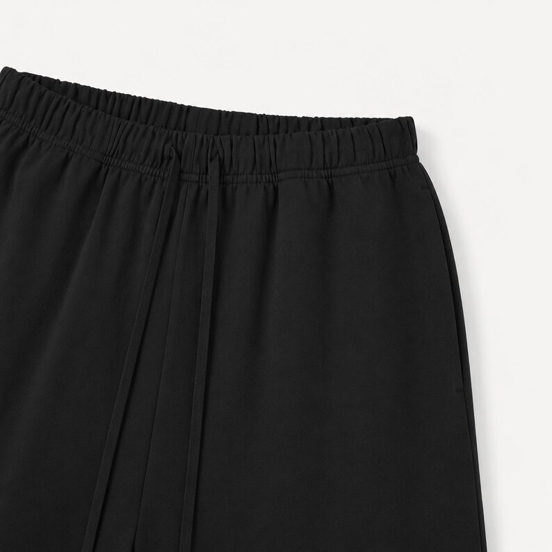 May include: Close-up of black trousers featuring an elasticated waistband and a drawstring. The material looks smooth and opaque. These trousers are likely part of a casual or sportswear collection.