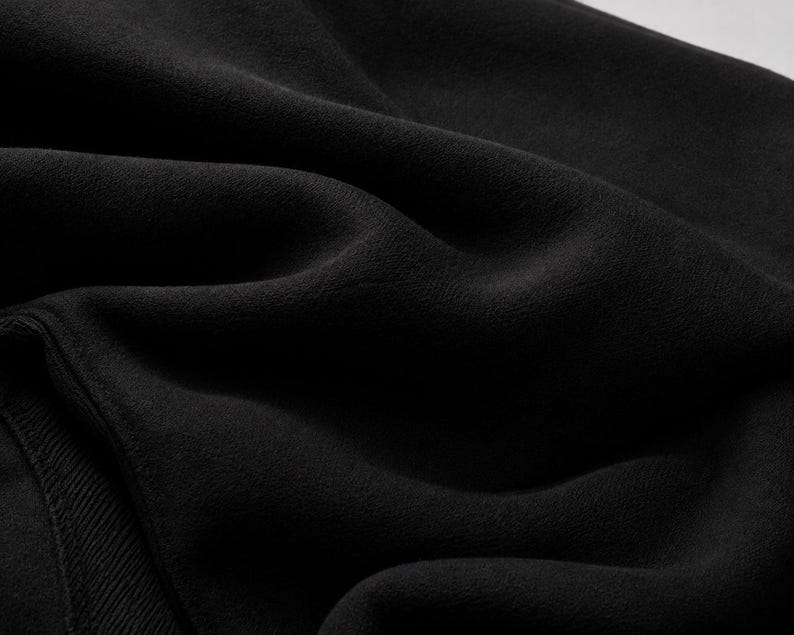 Puede incluir: Una tela negra s&oacute;lida con una textura suave y mate, con pliegues visibles que crean sombras y reflejos profundos en la superficie. El material presenta un sutil detalle de punto acanalado en el borde inferior izquierdo, de tono oscuro.