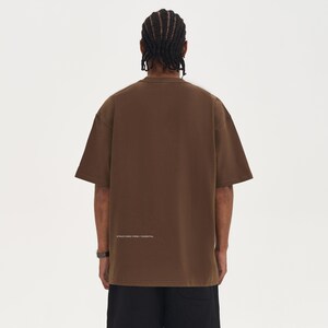 May include: A brown short-sleeve t-shirt with the text "STRUCTURED FORM / ESSENTIAL" printed in white near the bottom. The shirt has a crew neck and a relaxed fit. The person is wearing black shorts and a watch.