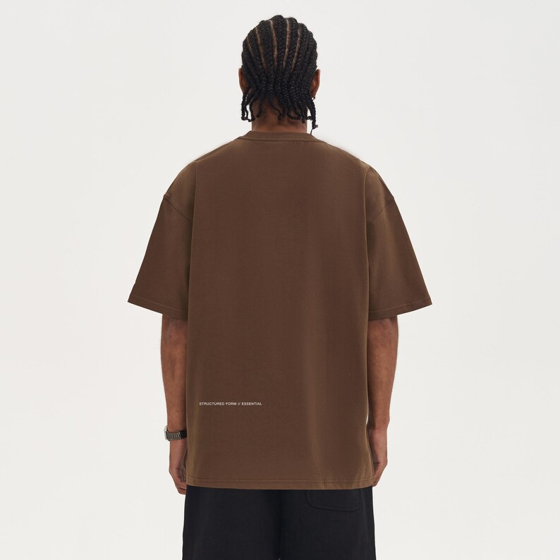 May include: A brown short-sleeve t-shirt with the text "STRUCTURED FORM / ESSENTIAL" printed in white near the bottom. The shirt has a crew neck and a relaxed fit. The person is wearing black shorts and a watch.