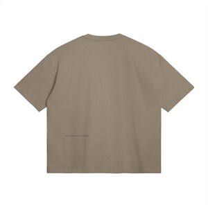 May include: A taupe-colored, short-sleeved t-shirt. The back of the shirt features a small, printed text near the bottom. The shirt has a relaxed fit and a crew neck.