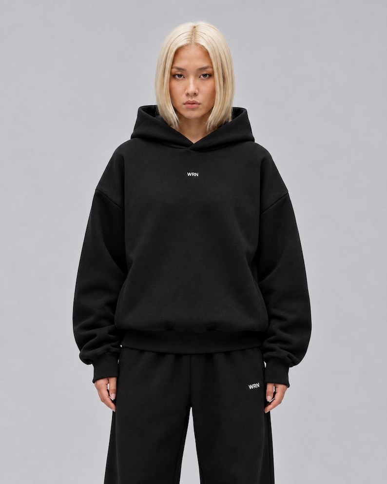 A black long-sleeve hooded sweatshirt featuring a hood, ribbed cuffs, and a ribbed hem. The text WRN is printed in small white capital letters on the centre of the chest. The garment has a relaxed fit with dropped shoulders.
