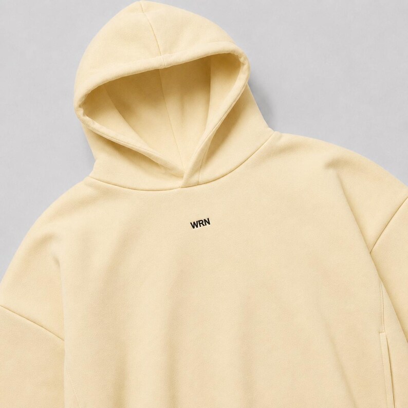 May include: A pale yellow hooded sweatshirt featuring a drawstring-free hood and long sleeves. The centre of the chest displays the text WRN printed in a small, bold, black sans-serif font. The garment has a smooth, solid-coloured fabric finish.