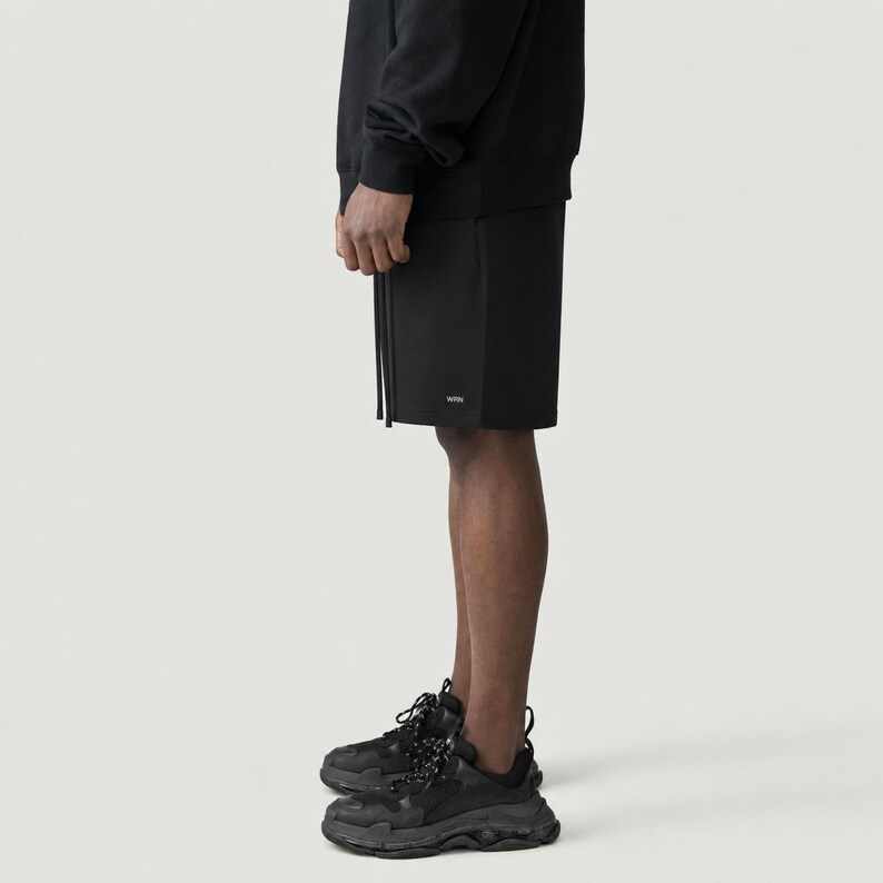 May include: Black shorts with a drawstring and the word "WRN" printed on the leg. The shorts are paired with black trainers with thick soles. The person is wearing a black sweatshirt.