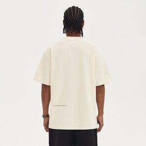 May include: Cream-colored short-sleeve t-shirt with the text "STRUCTURED FORM + ESSENTIAL" printed in black near the bottom hem. The shirt is worn by a person with black pants and a black watch.