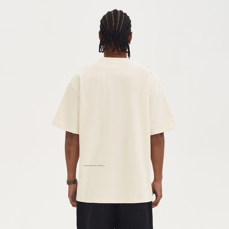 May include: Cream-colored short-sleeve t-shirt with the text "STRUCTURED FORM + ESSENTIAL" printed in black near the bottom hem. The shirt is worn by a person with black pants and a black watch.