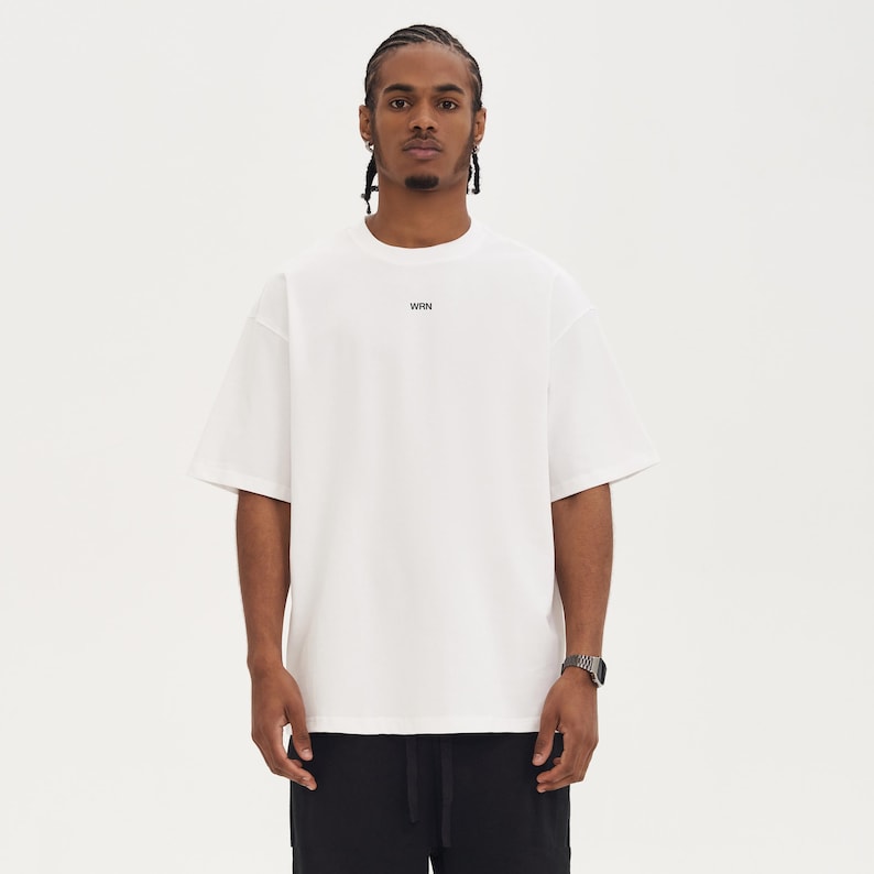 May include: A white t-shirt with short sleeves and a crew neck. The word "WRN" is printed in black on the chest. The model is wearing a watch and black pants. The background is plain white.