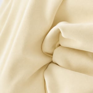 May include: A solid light cream-coloured sweatshirt featuring a smooth, matte fabric texture with visible folds and creases. The garment includes a ribbed hem detail at the bottom, showcasing a consistent, neutral tone throughout the entire piece.