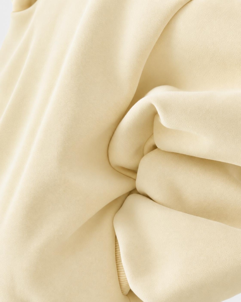 May include: A solid light cream-coloured sweatshirt featuring a smooth, matte fabric texture with visible folds and creases. The garment includes a ribbed hem detail at the bottom, showcasing a consistent, neutral tone throughout the entire piece.