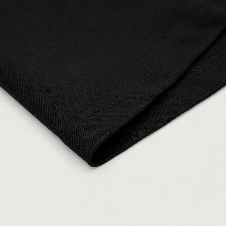 May include: Close-up of a section of black fabric, possibly a garment. The fabric has a smooth texture and is folded over, revealing a seam with visible stitching. The image is well-lit, highlighting the details of the material.