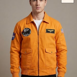 May include: Orange bomber jacket with a zip fastening, two front pockets, and patches. The jacket features a US flag patch, a winged emblem patch, and a patch with the text "US-AH PROJECT MARY". The jacket has ribbed cuffs and a collar.