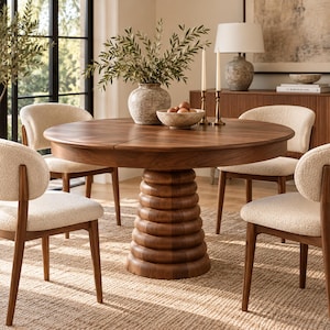 Può includere: Un tavolo da pranzo rotondo in legno con finitura marrone scuro e una base a piedistallo circolare a gradini. Il tavolo è circondato da quattro sedie con rivestimento testurizzato color crema chiaro e telai in legno scuro con schienali curvi.