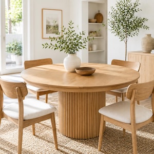 May include: A round light oak dining table with a fluted cylindrical pedestal base, accompanied by four wooden chairs featuring light-colored upholstered seats. A white ceramic vase with green foliage sits in the center of the table.