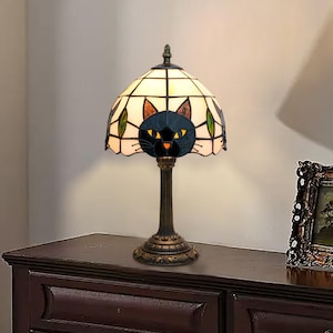 Könnte beinhalten: Tischlampe aus Buntglas mit einem schwarzen Katzenmotiv. Der cremefarbene Lampenschirm zeigt ein schwarzes Katzengesicht, gelbe Augen und eine orangefarbene Nase. Der Sockel ist dunkelbraun mit einem verzierten Design. Die Lampe ist ca. 46 cm hoch.