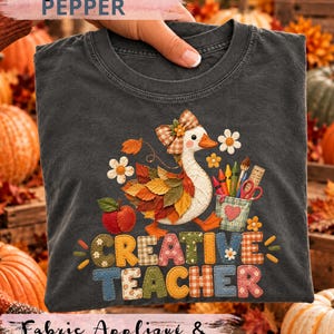 Puede incluir: Una camiseta gris carbón con un gráfico de un ganso blanco con alas de hojas de otoño, un lazo a cuadros y un portalápices. El texto CREATIVE TEACHER aparece en letras de estilo patchwork coloridas y estampadas junto a una manzana.