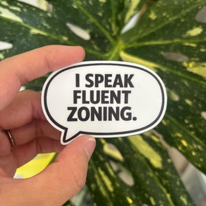 May include: A white speech bubble sticker with a black border featuring the black text I SPEAK FLUENT ZONING. The sticker has a smooth, matte finish and is shaped like a classic comic book dialogue balloon with a small tail pointing downward.