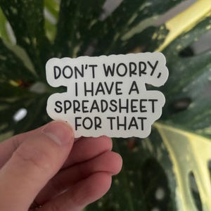 May include: A white sticker with black text that reads "DON'T WORRY, I HAVE A SPREADSHEET FOR THAT." The sticker is held in front of a green and yellow leafy background.