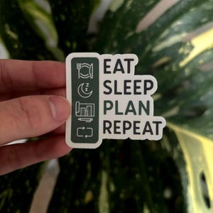 Puede incluir: Una pegatina blanca con las palabras "EAT SLEEP PLAN REPEAT" en negro. La pegatina tiene un borde verde con iconos que representan comer, dormir, planificar y repetir. La pegatina se sostiene frente a una hoja verde y amarilla.