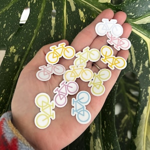May include: A selection of bicycle stickers in pastel shades of yellow, pink, blue, and white. The stickers are held in a hand, with a green and yellow leaf in the background. The stickers are small and feature a simple bicycle design.