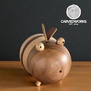 Könnte beinhalten: Eine handgefertigte Holzbiene mit rundem Körper und Streifenmuster. Die Biene hat kleine Holzaugen, einen lederartigen Flügel und ein kleines Loch. Das Logo "CARVEDWORKS EST. 2025" befindet sich im Hintergrund. Die Biene steht auf einer Holzoberfläche.