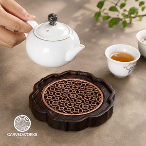 Könnte beinhalten: Eine weiße Keramik-Teekanne mit silberfarbenem Filigran-Deckelknopf, eine kleine weiße Teetasse mit bernsteinfarbener Flüssigkeit und ein dunkelbraunes Holz-Teetablett mit kupferfarbenem Metalleinsatz und kompliziertem geometrischem Gittermuster.
