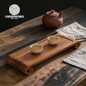May include: A wooden tea tray with two small teacups filled with tea. A brown teapot sits in the background. The tray has a carved design and the text "CARVEDWORKS EST. 2025" in the upper left corner.
