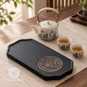 Könnte beinhalten: Ein schwarzes, rechteckiges Teetablett mit einem runden, bronzefarbenen Einsatz, der ein detailliertes Drachenrelief zeigt. Das Set enthält eine helle Keramik-Teekanne mit Landschaftsmotiv und Seilgriff sowie zwei passende Keramik-Teetassen.