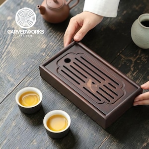 以下が含まれることがあります： 彫刻された鳥のデザインとスロット付きの表面を備えた、濃い茶色の木製ティートレイ。 2つの小さな白いティーカップにはお茶が入っています。 茶色のティーポットと小さなセラミック花瓶も表示されます。