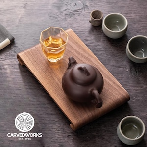 Könnte beinhalten: Ein hölzernes Teetablett mit einer braunen Teekanne, einem facettierten Glas Tee und mehreren kleinen Keramikbechern. Das Tablett hat gebogene Kanten. Das Logo "CARVEDWORKS EST. 2025" befindet sich in der unteren linken Ecke.