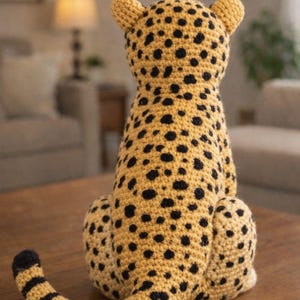 K&ouml;nnte beinhalten: Handgefertigte H&auml;kelfigur eines Geparden. Das Pl&uuml;schtier ist haupts&auml;chlich beige mit schwarzen Flecken, kleinen Ohren und einem gestreiften Schwanz. Der Gepard sitzt auf einem Holztisch. Der Hintergrund ist unscharf.