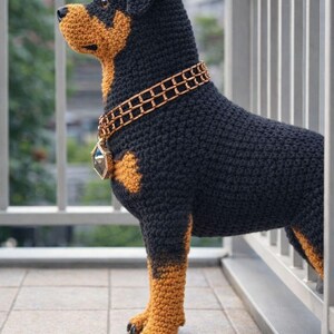 Puede incluir: Escultura de perro Rottweiler de ganchillo, predominantemente negro con marcas marrones, de pie sobre una superficie embaldosada. Presenta un collar decorativo de color dorado. La escultura hecha a mano es detallada y realista, mostrando la textura del hilo.