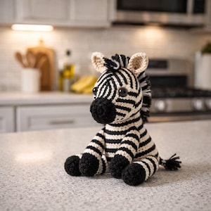 Może przedstawiać: Ręcznie robiona szydełkowa pluszowa zebra. Zebra ma czarno-białe paski, czarny nos i czarne kopyta. Uszy są beżowe. Zabawka siedzi na blacie.