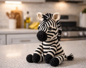 May include: A handmade crochet zebra plush toy. The zebra features black and white stripes, a black nose, and black hooves. The ears are beige. The toy is sitting on a countertop.