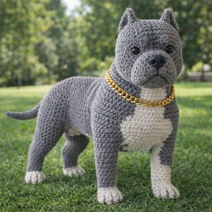 Op de afbeelding: Gehaakte hondensculptuur in grijstinten en wit, met een goudkleurige kettinghalsband. De hond staat op groen gras, met een vage achtergrond van bomen. De handgemaakte hond heeft een realistisch uiterlijk.