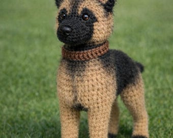 May include: A crocheted German Shepherd dog figurine. The dog is tan and black, with a brown collar. The dog is standing on green grass.