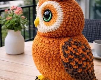 May include: A handmade orange crochet owl figurine with large, expressive eyes. The owl has white and brown accents, a yellow beak, and yellow feet with black claws. It is sitting on a wooden table, showcasing its detailed craftsmanship.