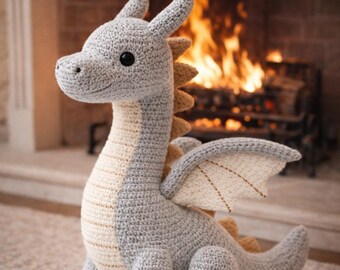 May include: A crocheted dragon toy in shades of gray, beige, and tan. The dragon has large black eyes, a long neck, and wings. The background shows a fireplace with a warm fire burning.