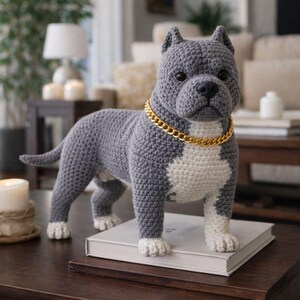 May include: A crocheted dog figurine in shades of gray and white, with a gold-colored chain collar. The dog is standing on a book, with its paws and chest in white. The background includes a table, candles, and a lamp.