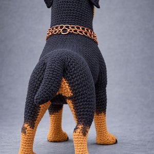Puede incluir: Figura de perro hecha a ganchillo, vista desde atr&aacute;s. El perro es principalmente negro con marcas marrones en las patas. Lleva un collar de cadena color cobre. El trabajo de ganchillo muestra la textura de las puntadas.