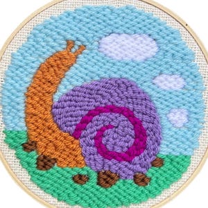 Op de afbeelding: Een handgemaakte borduurring met een slakkenontwerp. De slak heeft een paars huisje met een roze spiraal, een oranje lichaam en staat tegen een blauwe lucht met witte wolken en groen gras. Het kunstwerk is omlijst in een houten ring.