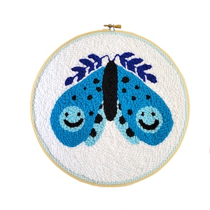 Op de afbeelding: Een blauwe en zwarte vlinder met lachende ogen is geborduurd op een wit canvas in een houten borduurring.
